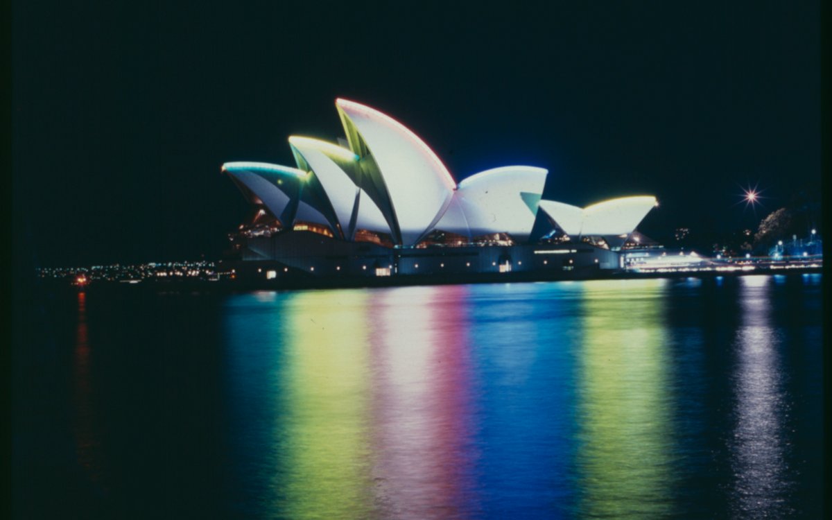 A nighttime photograph of the Sydney Opera House. The sails of the Opera House are lit up in different colours that are reflecting green, yellow, pink, blue and chartreuse into the water of the harbour.