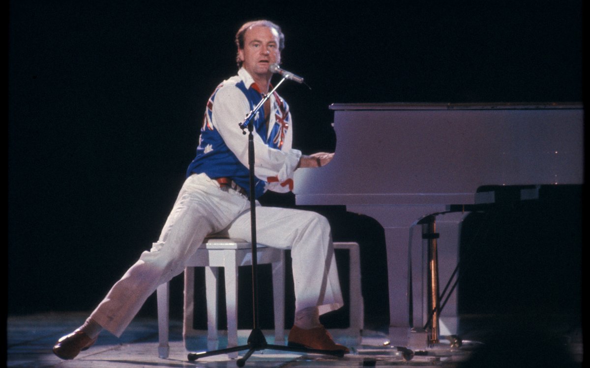 A man wearing white pants, shirt and an Australian flag vest plays a white grand piano on stage
