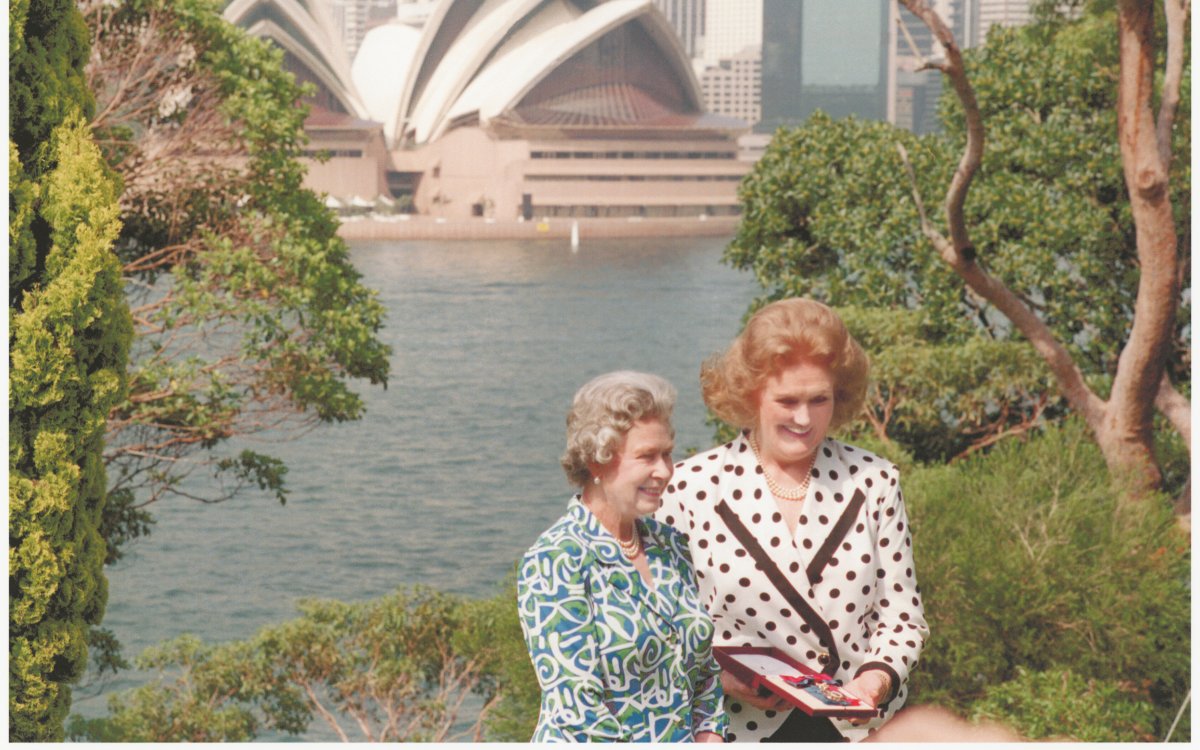 Two women stand among trees with the Sydney Opera House in the background. One lady wears a white and black polka dot blazer and has red hair. The other wears a green, white and blue patterned dress. She has grey hair. Both are smiling
