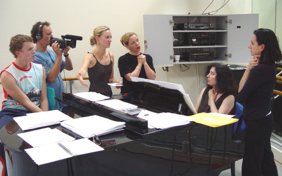 Four women and two men stand around a black baby grand piano in a studio. One man is filming the pianist and is wearing headphones. The group are mid-conversation.
