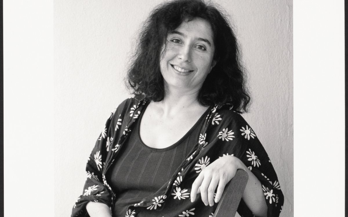 A black and white portrait of a woman sitting sideways in a chair. She has one arm over the back of the chair and the other in her lap. She has black wavy hair. She is wearing a black coat with daisies and a black undershirt.