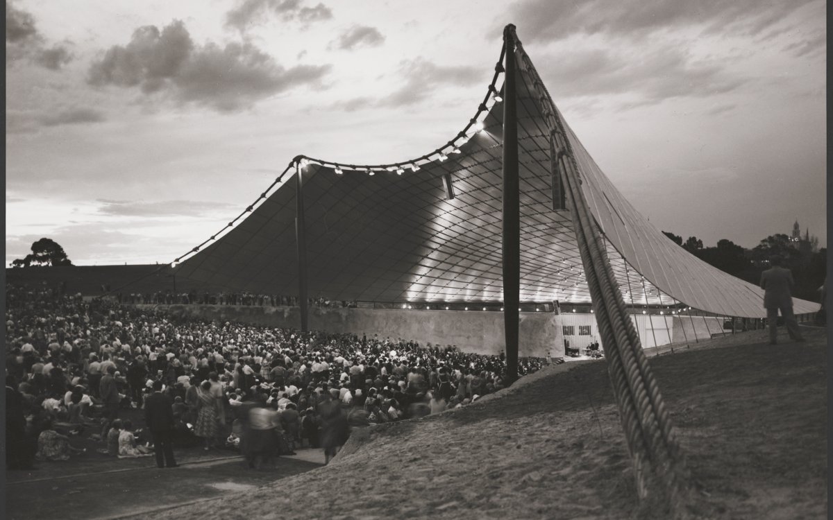 A sepia photograph of the Sydney Myer Music Bowl. A large trianglular sail like building with a stage. A large crowd are gathered watching the stage.