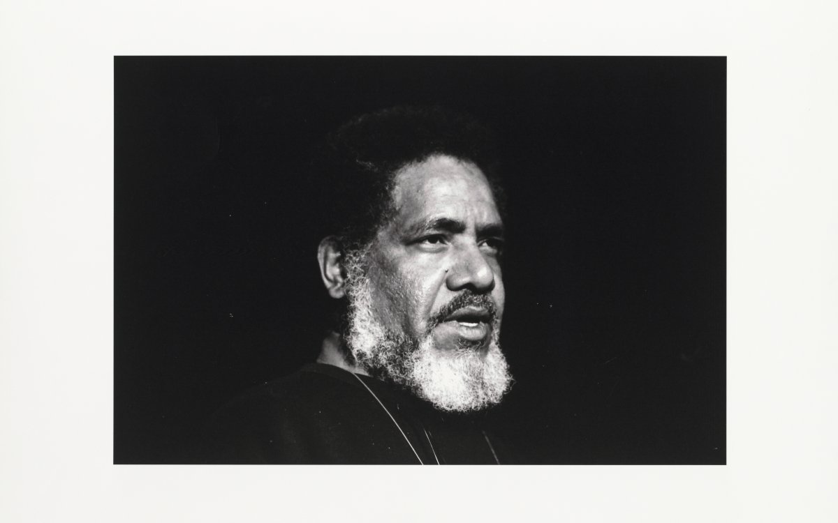 A black and white photograph of a man standing in front of a black background. He has a white beard and black hair. He is staring off camera mid-speech. The picture is surrounded by a thick white frame