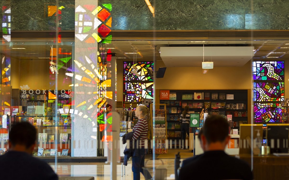 A colour photograph of the front of the National Library Bookshop, showing people sitting and walking in the foreground and the Bookshop in the background
