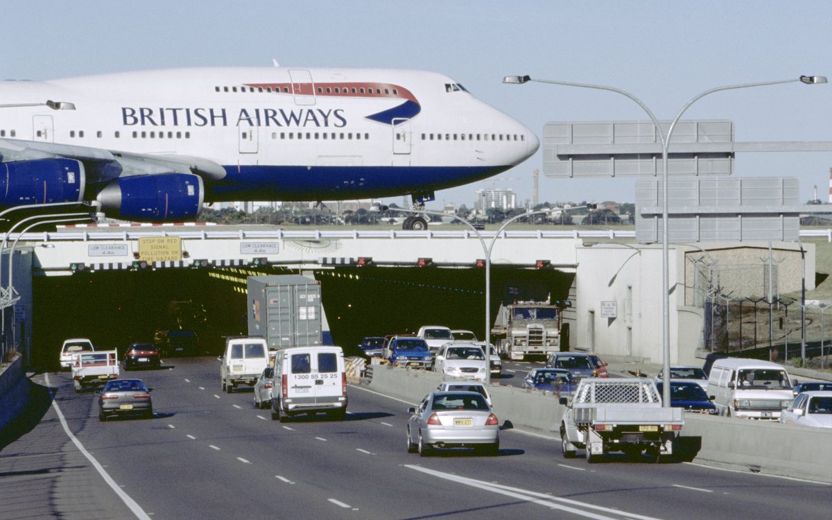 A British Airways Boeing 747 Jumbo Jet taxis across the runway bridge over a highway tunnel. Cars are entering and exiting the tunnel