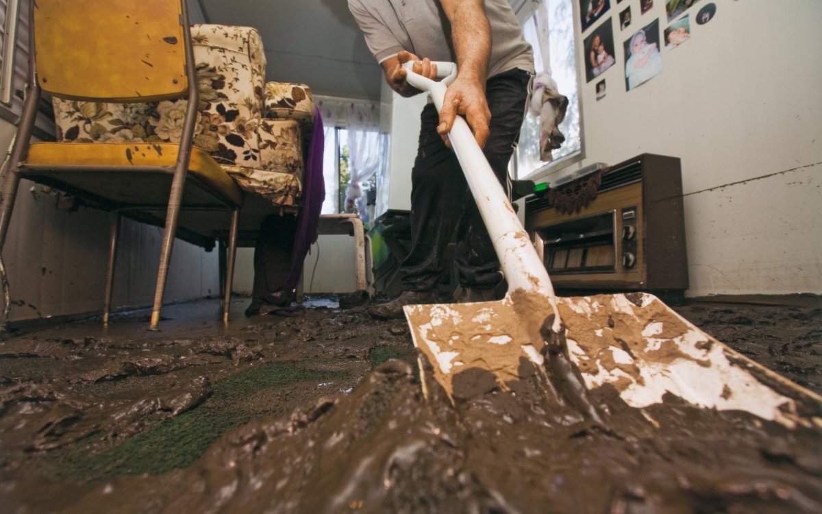 An interior shot of a room. A person is scooping up silt and mud with a shovel. The furniture is covered in mud.