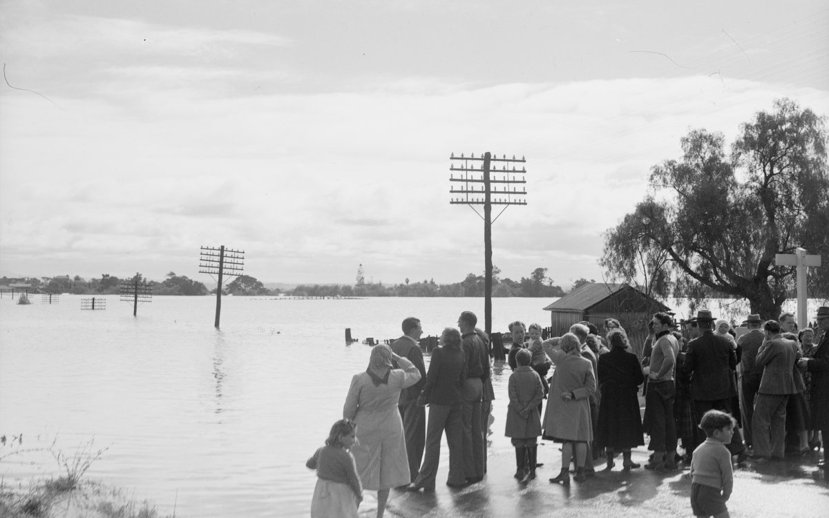 A large group of people stand on a road that is cut off by a swollen flooded river. Telegraph poles are submerged up to the head and trail off into the water.