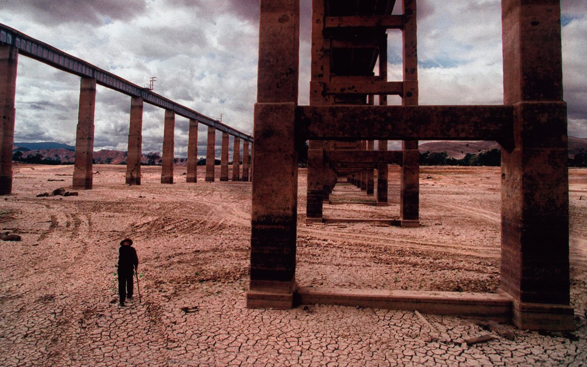 A man with a walking stick walks below the pylons of a huge bridge that have surfaced after a drought. The river bottom is cracked and crazed with the drying of the mud.