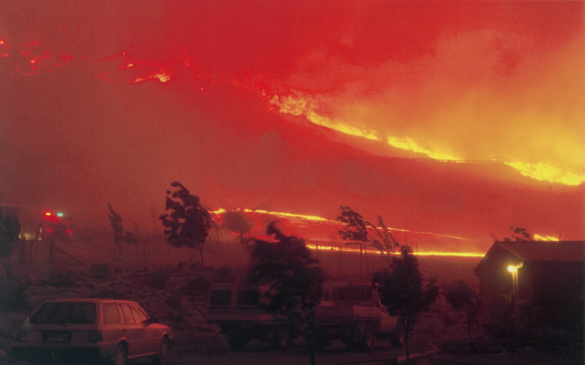 A scene of a hillside completely ablaze. A large fire front is moving up the hillside. The image is blanketed with smoke. The lights of a fire engine can be seen through the smoke. A station wagon is parked in the forground.