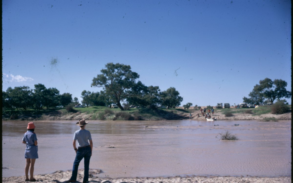 Two people stand on the bank of a swollen river in full flood. The water is turgid and brown. In the distance, a group of people stand on the opposite bank.