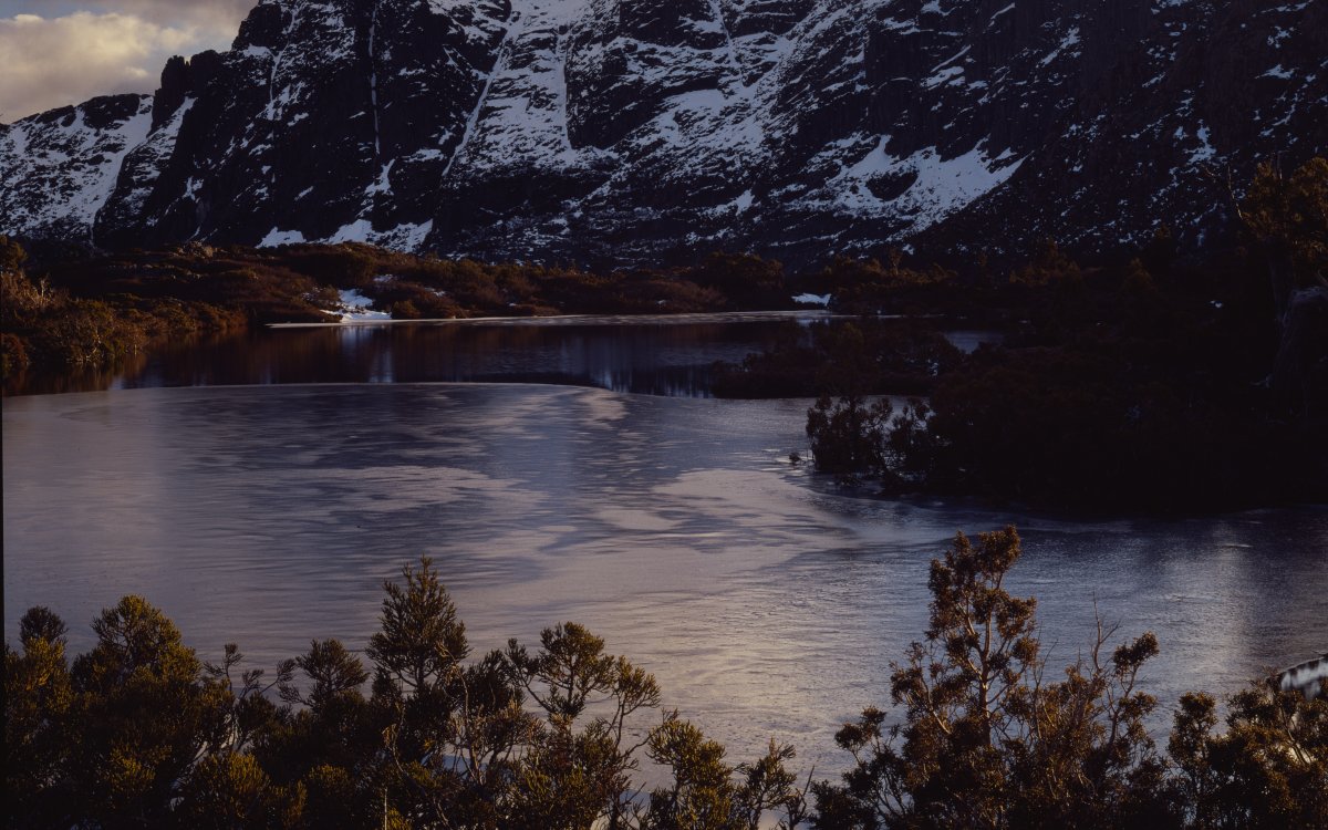 A landscape of a mountain lake scene. The craggy mountains are dusted with snow. The lake is surrounded by trees with yellow leaves.