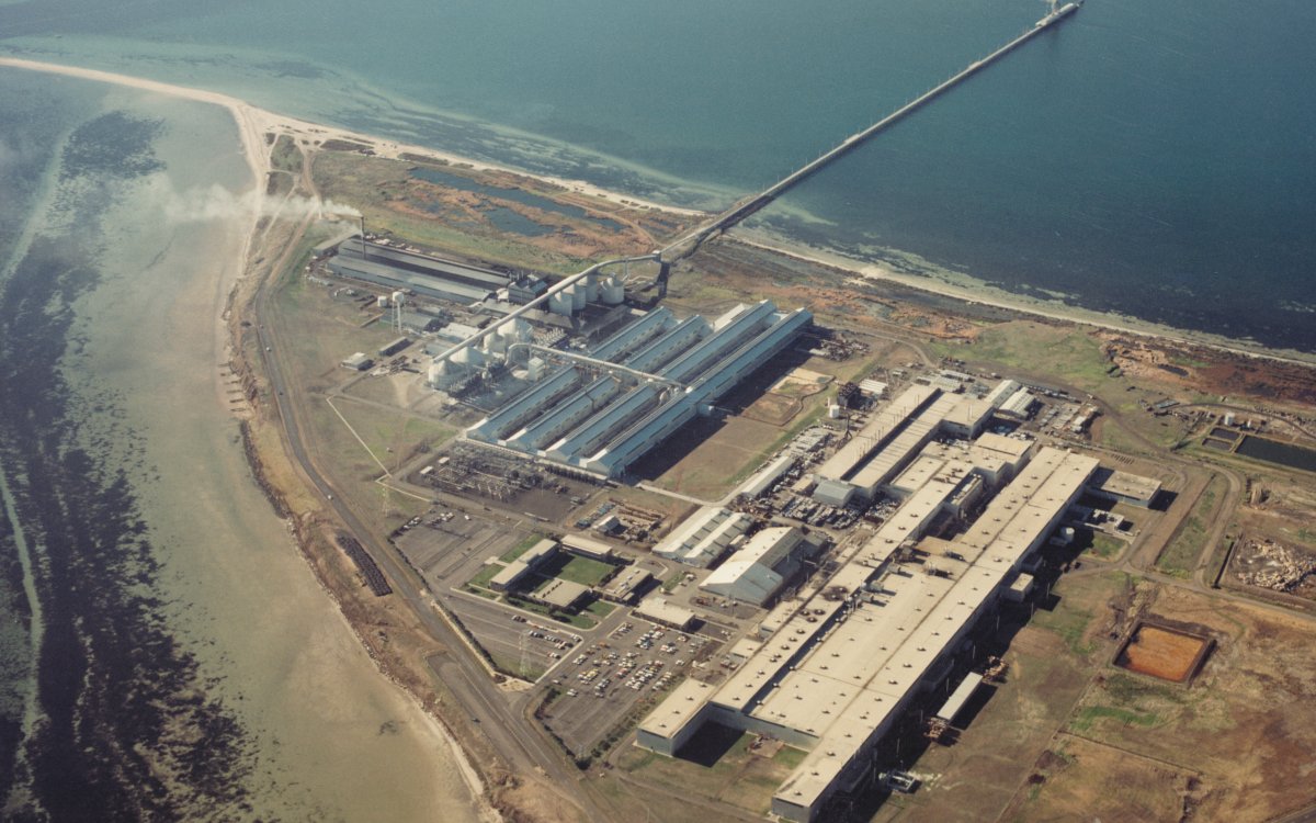 An aerial shot of a huge factory on a point surrounded by ocean. The factory is made up of several large sheds and warehouses. A long pier runs out into deep water.