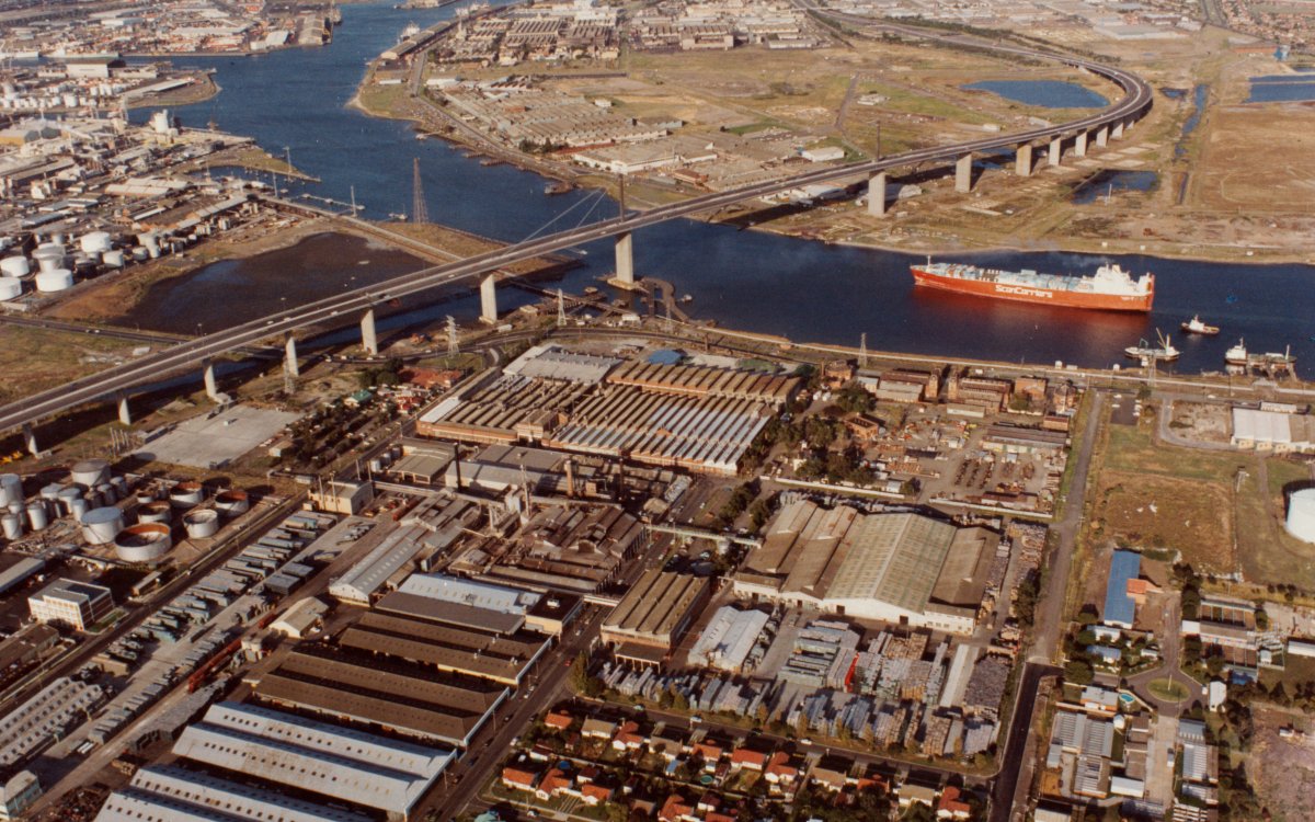 An aerial shot of a city port industrial district. A large river winds through the middle. A large ship is on the river about to sail under a bridge.