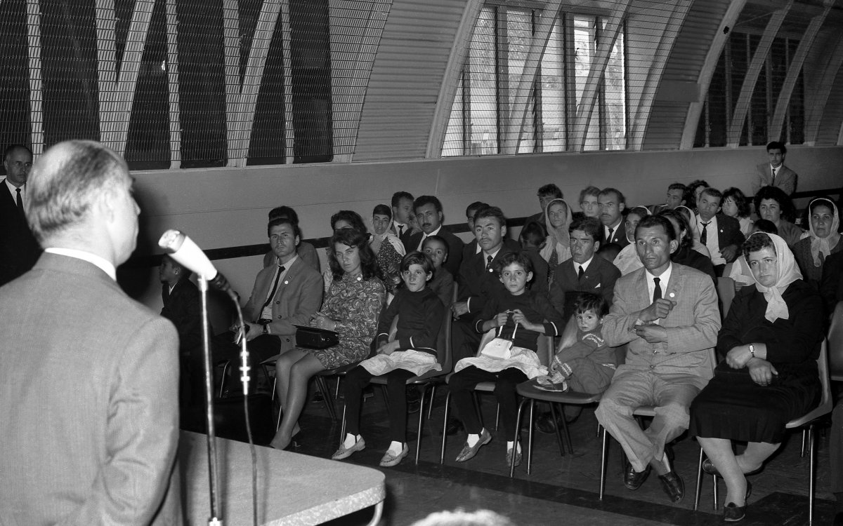 A photograph of a large group of people seated in an auditorium. A balding man is standing at a microphone addressing the audience.