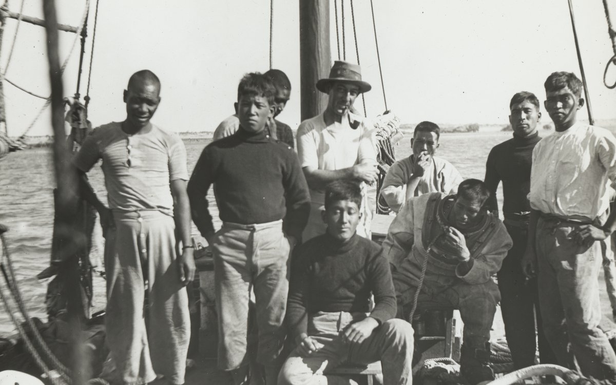 Nine men stand on the deck of a small boat. One is wearing a hat, the others are not. Three men are sitting. In the background the ocean can be seen.