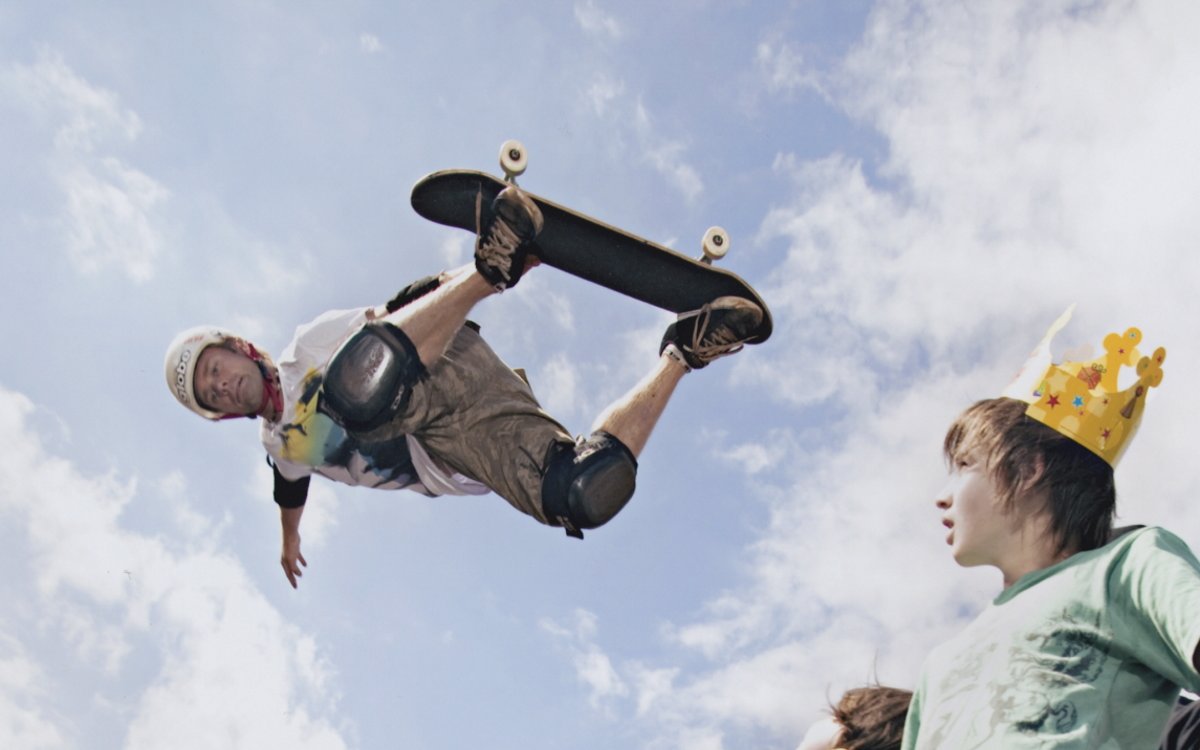 A person on a skateboard is in the air. Two people in the bottom right hand corner are looking up at him. One of them is wearing a cardboard crown. The background is a blue sky with white clouds.