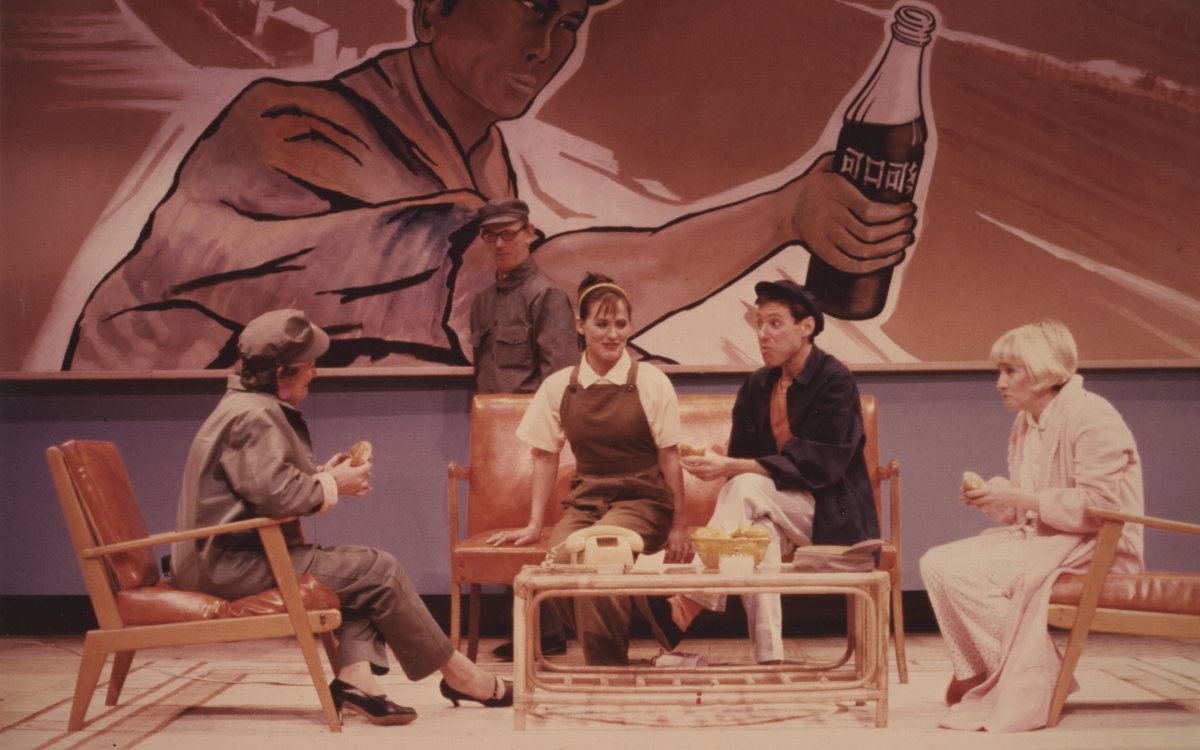 Four people sitting on couches and armchairs around a coffee table. There is a large artwork on the wall behind them of a man in a hat holding a bottle of softdrink.