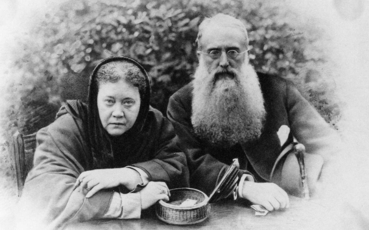 A black and white image of a woman and a man sitting down at a table. There is some kind of shrubbery in the background. The woman is wearing a head scarf covering. The man has a long beard and wears glasses. He is holding a cane and a hat. On the table is an small wicker basket that has been opened, and there is something indistinguishable inside it. At the bottom of the image, under each person, are their signatures.