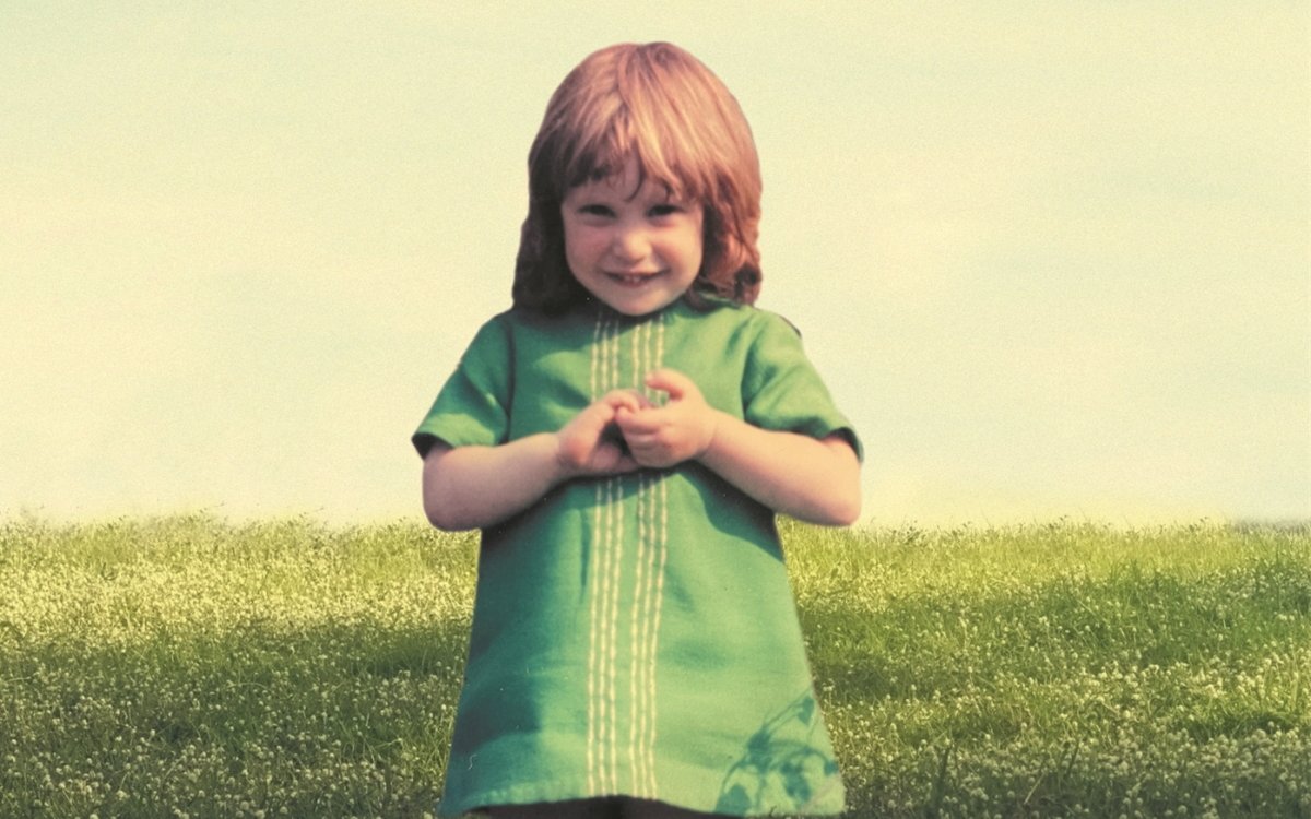 An image of a little girl in a green dress. She is smiling and has her hands held in front of her, clasping them together. In the background is green grass and a greenish-yellow sky.