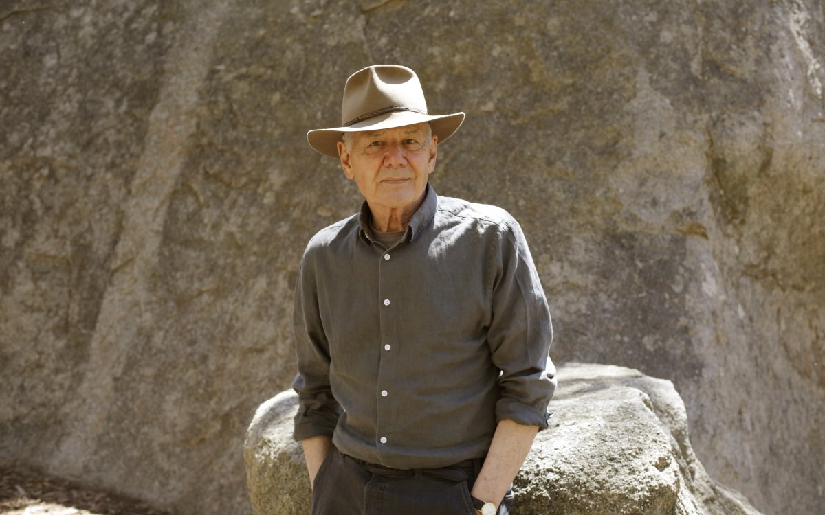 A portrait image of a person. They are standing against a rock, with a rock face behind them. They are wearing a hat, a brown collard shirt and black pants. Their hands are in their pockets.