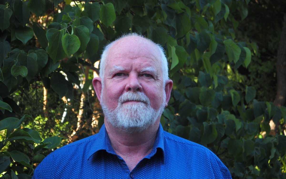 A man standing in front of green shrubbery wearing a blue collard shirt. He was white hair and facial hair.