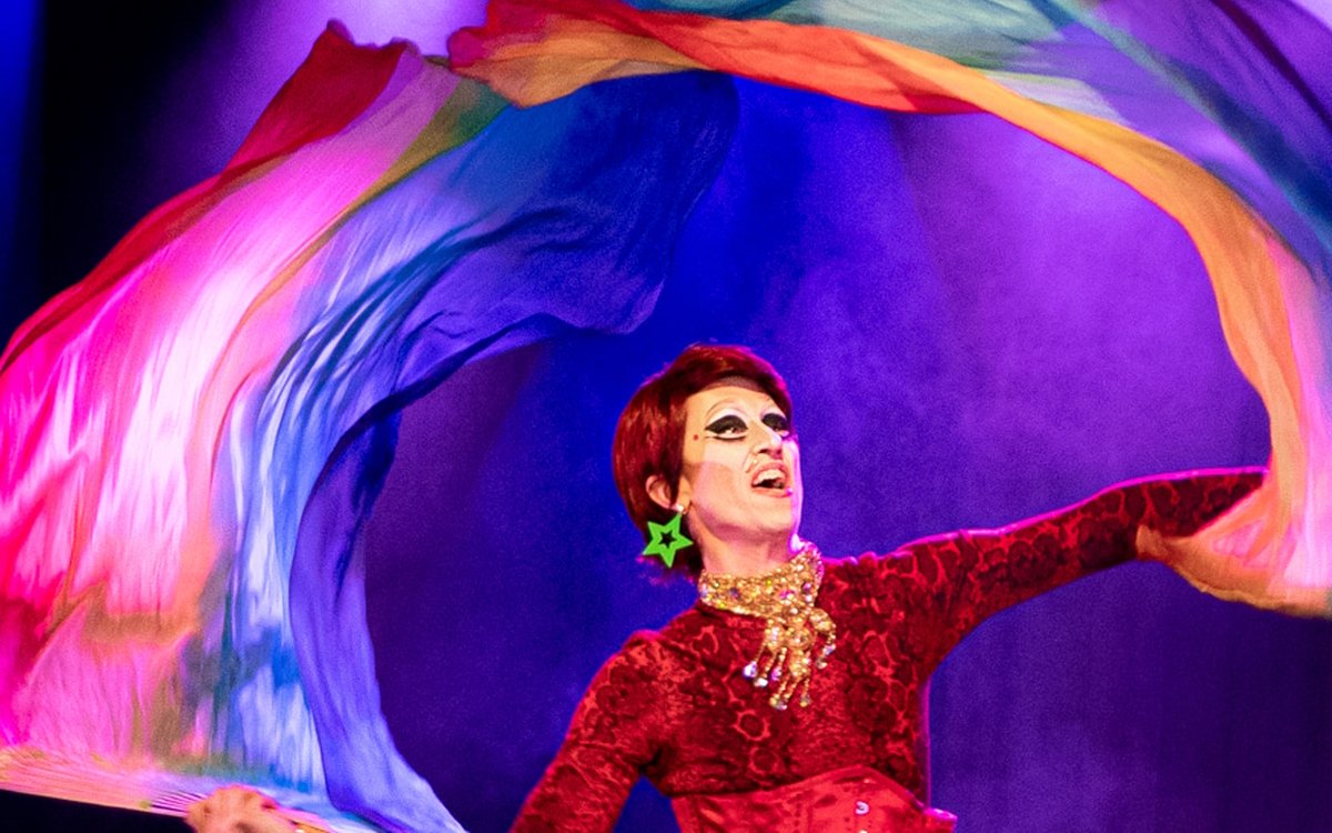 An image of a person with short hair in a red outfit fluttering a long rainbow silk piece of material above their head.