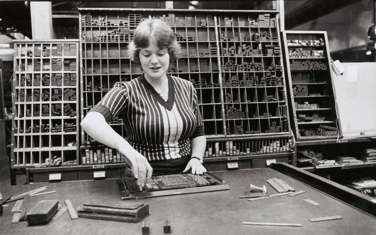 A black and white image of a woman at a desk setting type face into a frame. Behind her is a large rack full of letters