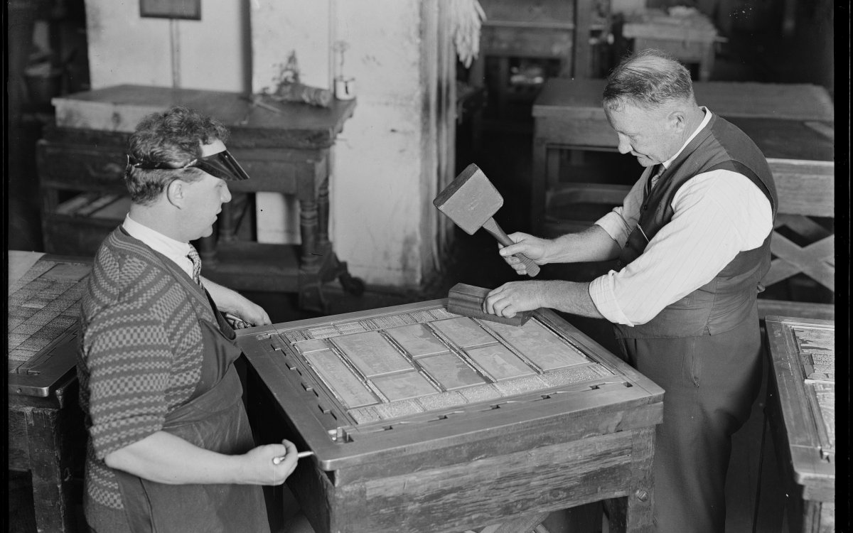 A black and white image of two men setting a page up with typeface. One man is setting the screws of the frame while another man makes  sure all the letters are set by hitting them with a wooden mallet.
