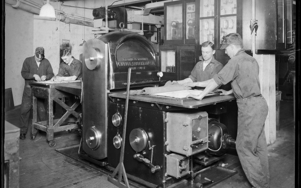 A black and white image of a group of men standing around a large industrial press.