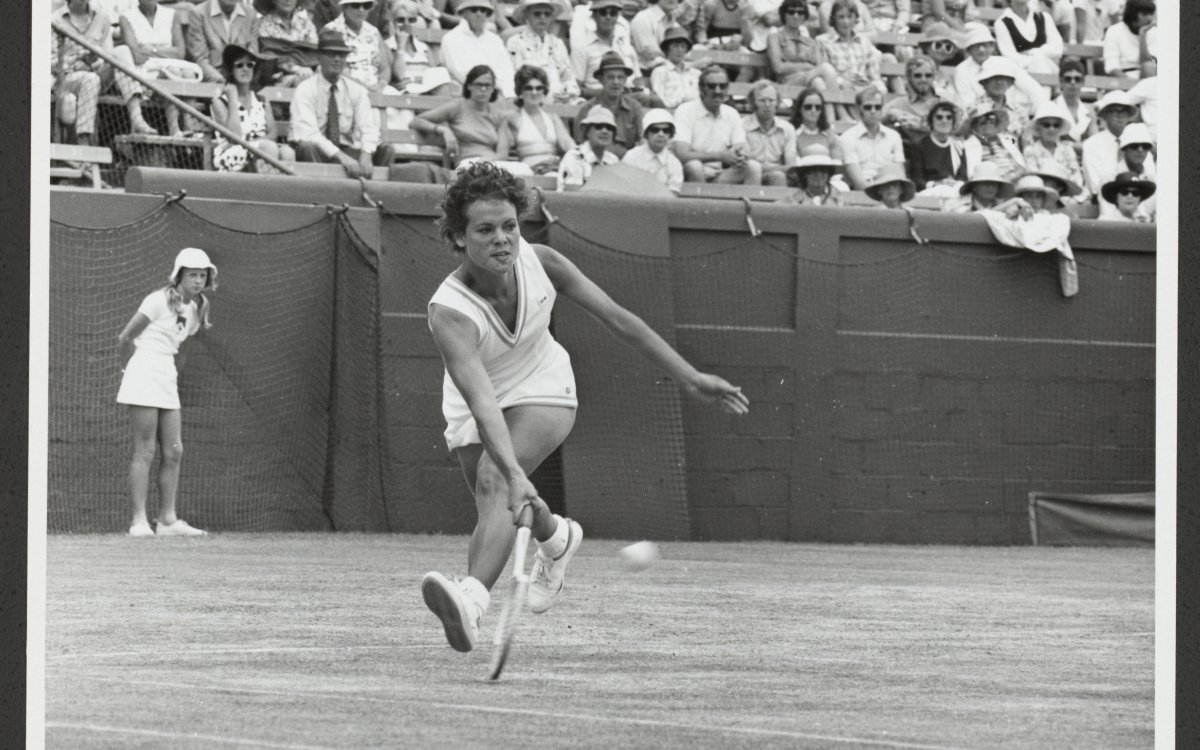 A black and white image of a woman wearing a white tennis dress lunging to hit a tennis ball. Behind her is a young woman in a white tennis dress and white bucket hat watching. There is a large crowd watching the game of tennis that is being played.