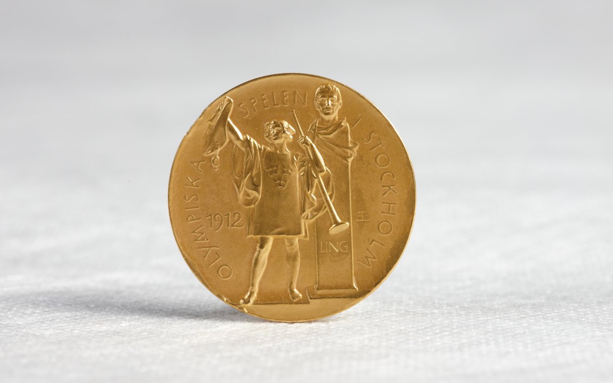 A gold medal standing upright on a white cotton surface. The medal shows a figure holding a trumped and a wine amphorae. Behind the figure is a bust on a stand. In raised lettering around the edge of the medal are the words "Olympiska Spelen Stockholm" The year "1912" is also present.