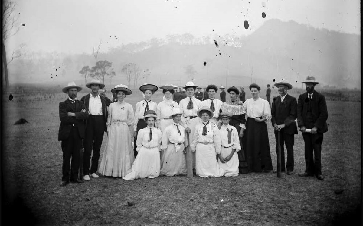 A black and white photo of 11 women standing in all white dresses flanked by 2 men on either side of them, dressed in black.