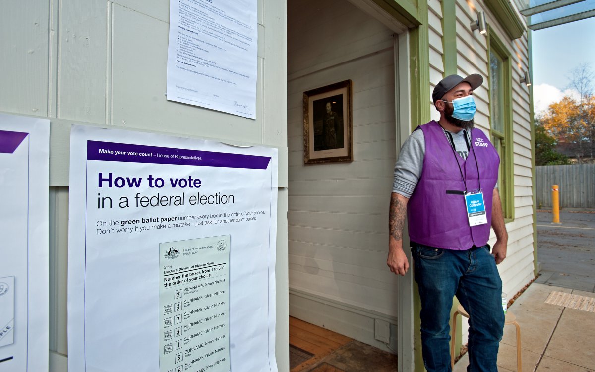A photograph of a man wearing a purple vest that says "AEC STAFF" and a blue medical facemask. He is standing in front of a building one which posters explaining how to vote in a federal election are hanging.