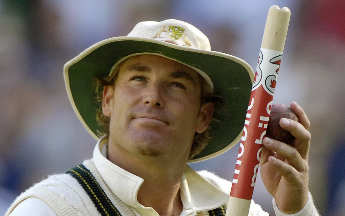 A man in cricket whites and a hat holding a cricket stump and looking upwards.