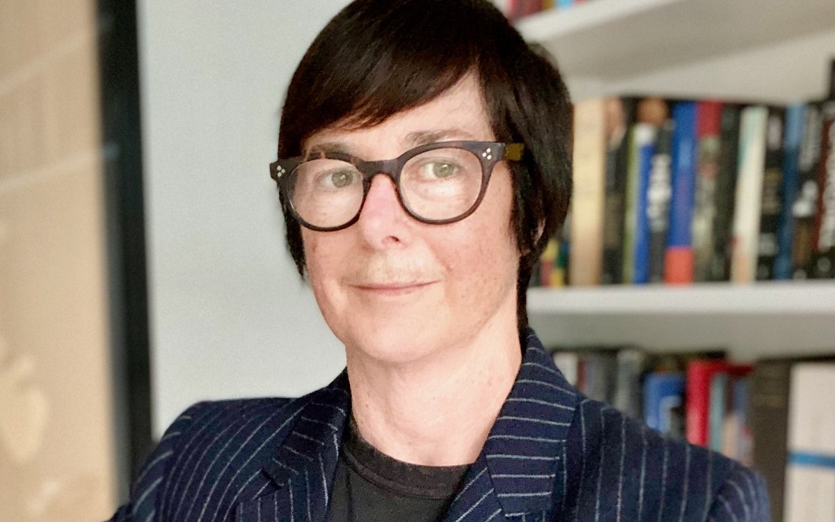 A portrait image of a person with short hair, wearing glasses and a dark blue striped blazer. They are standing in front of a bookshelf.