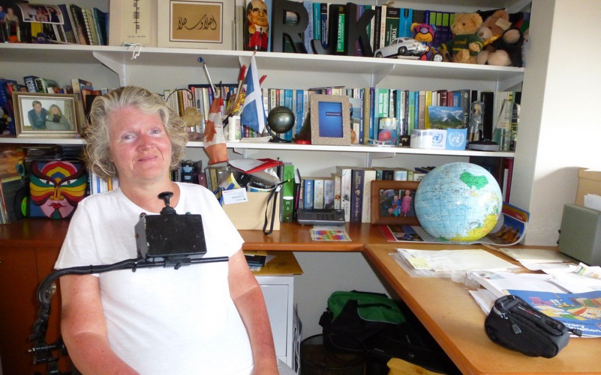 A photograph of a woman. She is seated in a wheelchair in front of a large bookcase and desk. She is wearing a white tshirt. The wheelchair has a control stick which sits just below her chin. She is smiling.