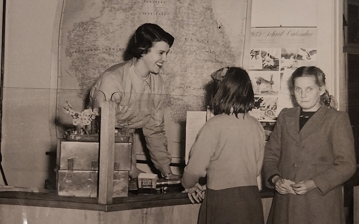 A sepia image of a lady behind a counter talking to two young girls who are standing in front of the counter.