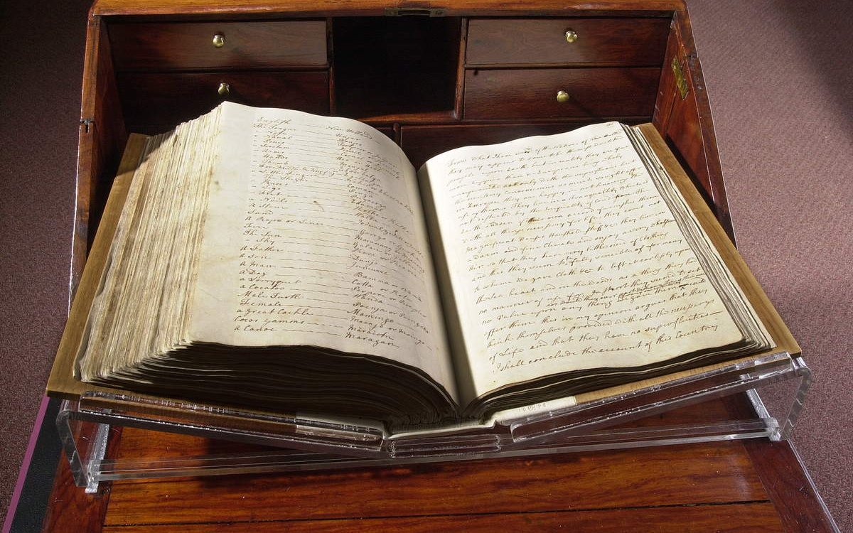 A large book open at a midway point. The paper is yellowed and stained. The writing is written in highly decorative cursive. The book is being held open on a stand which sits atop a richly varnished cabinet with draws and a fold out desk surface.