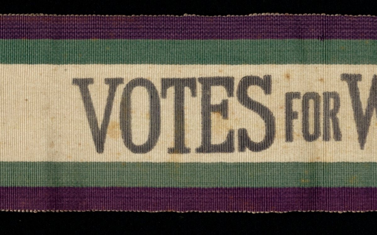 A fabric sash in purple, green and white with the words "Votes for Women"