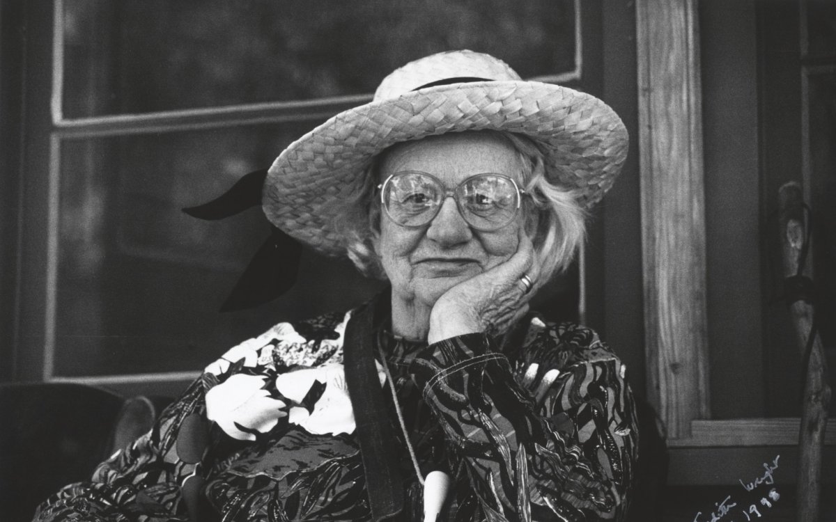 A black and white portrait photo of an old woman with white hair under a straw sunhat, wearing large reading glasses, and a long sleeved patterned dress, sits smiling at the camera with her hand resting, cupping the left side of her chin and face.