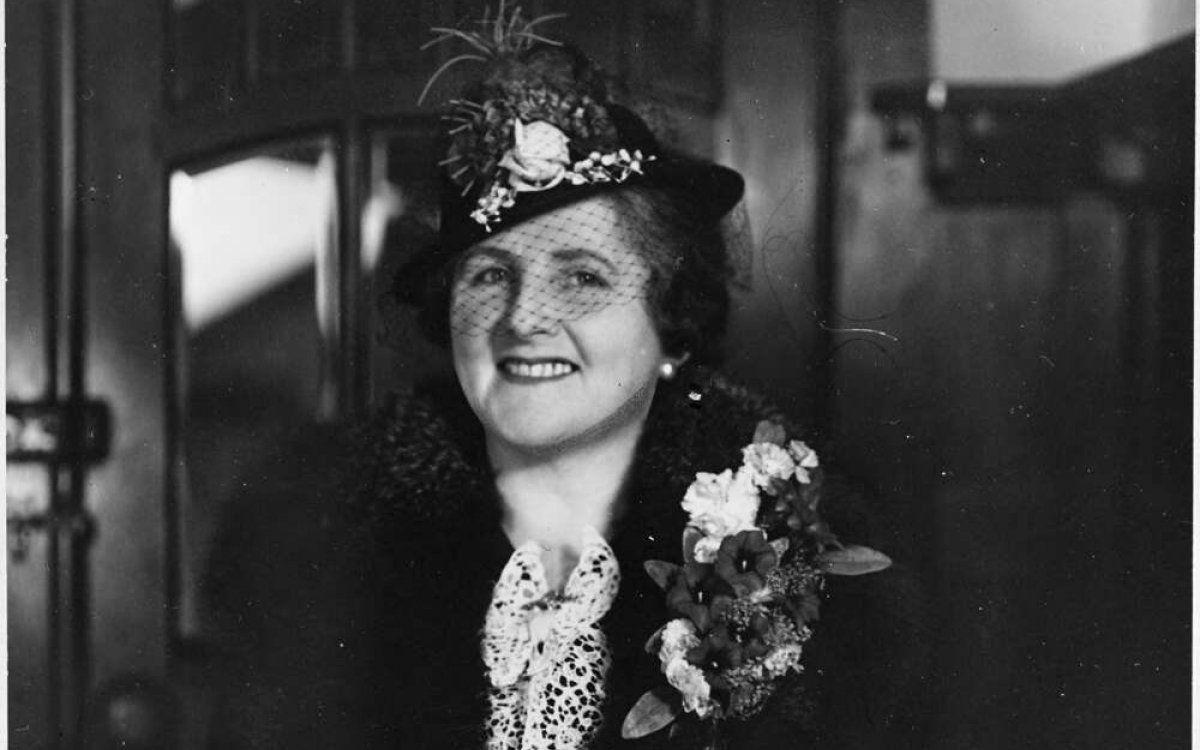 A black and white photograph of a woman. She is wearing dark clothes and a hat with a feather  plume. She is smiling broadly.