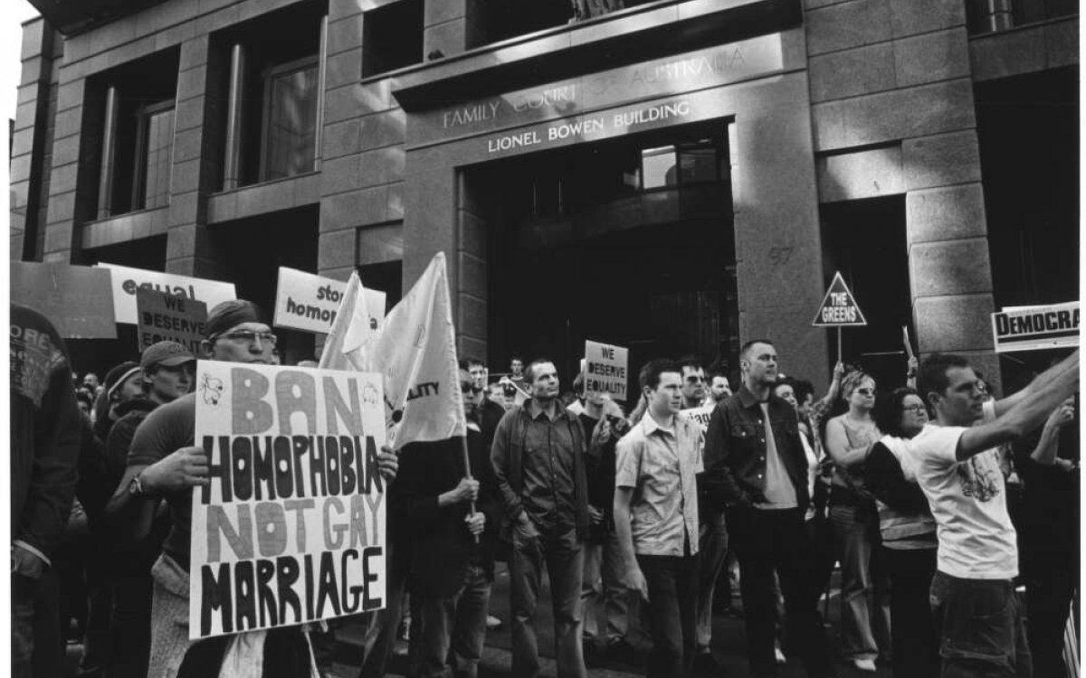 A black and white photo of a large number of people marching through city streets with signs supporting gay marriage. One sign says 'Ban homophobia, not gay marriage'.