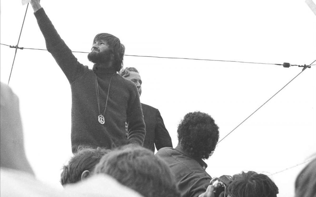 A black and white photo of a bearded man with collar length hair and a large metal peace medallion hanging down his front, holds a burning piece of paper up high, while several people watch and take photos.