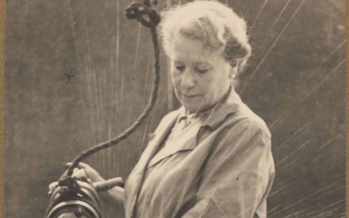 A sepia toned photograph of a woman standing at a workbench. She is holding an angle grinder. She is grinding an metal object on the workbench. Sparks shoot out from where the tool and the object are touching.