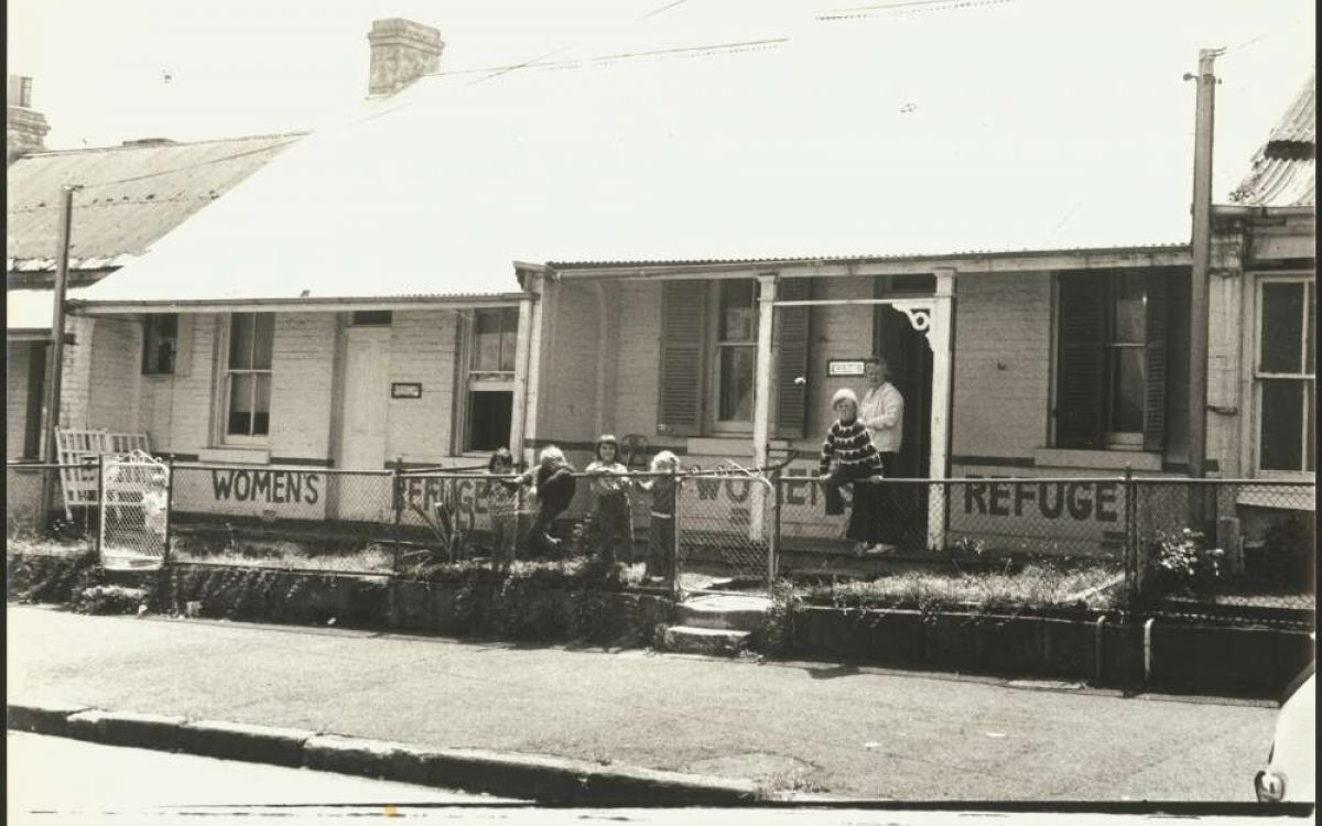 A black and white photograph of a row of houses. On the verandah of to adjacent houses aare the words "WOMENS REFUGE". A women and a group of children are standing next to and sitting on a the fence.