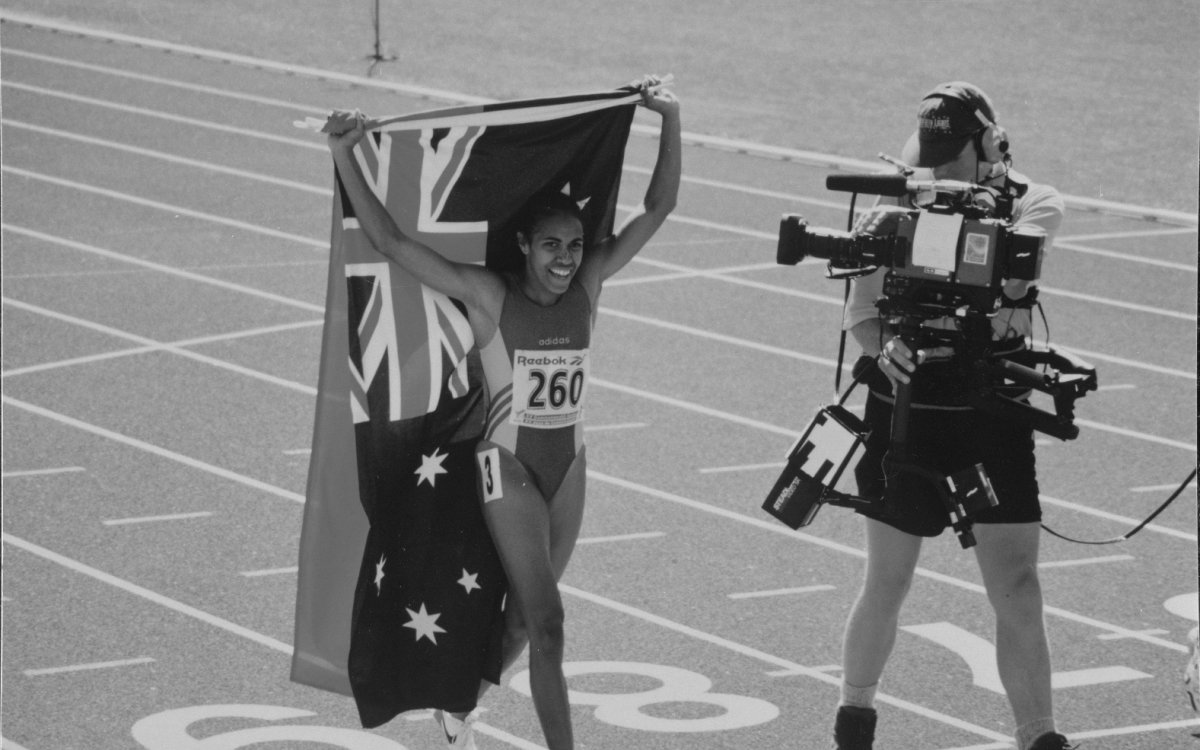 Black and white photo of Cathy Freeman carrying Australian and Indigenous flags with a cameraman filming alongside her