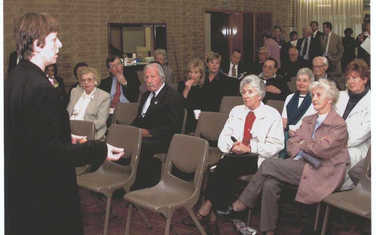 A photo of a woman standing and speaking to a room of seated and standing people.