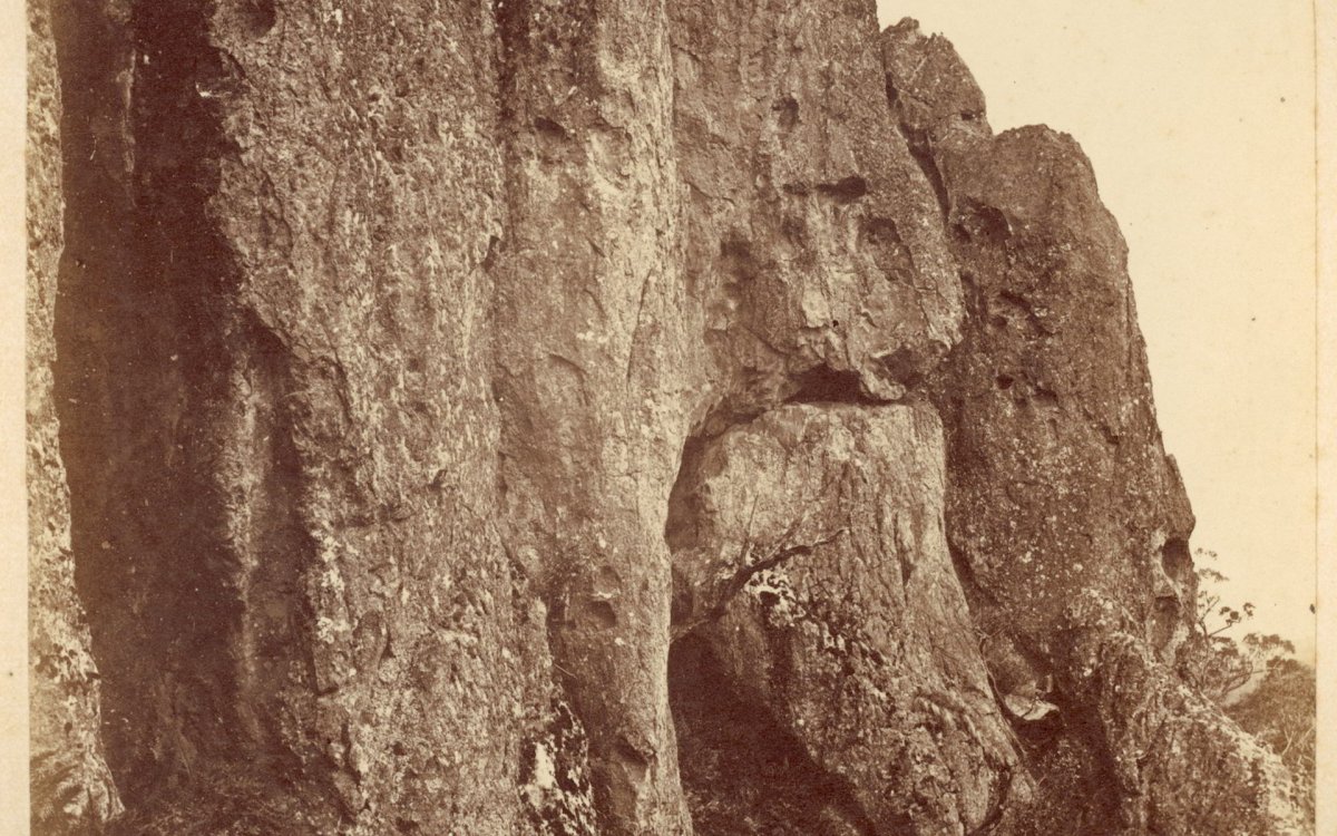 A sepia toned photo of a closeup of Hanging Rock, with words typed underneath saying ' View of the "Hanging Rock" near Mount  Macedon'