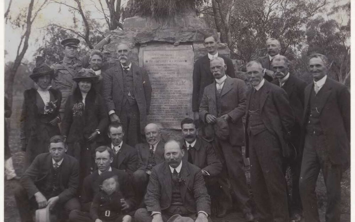 A black and white photo showing 15 men suits, including a man in an army uniform, 2 women and a child , gathered in front of a monument with a large plaque.