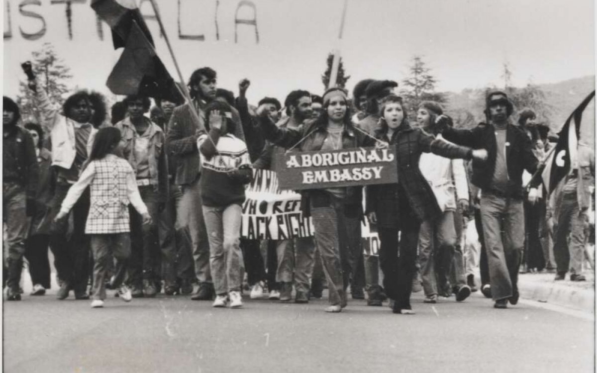 A black and white photo of a large group of people marching along a street, holding large flags and a sign saying 'Aboriginal Embassy'.